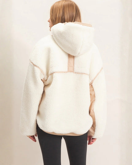 A woman viewed from behind wearing a cream-colored sherpa fleece hooded jacket with beige contrast panels and black leggings, set against a soft, light beige backdrop.