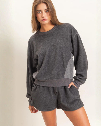A woman modeling a dark gray heathered crewneck sweatshirt and matching shorts set against a plain off-white studio background.