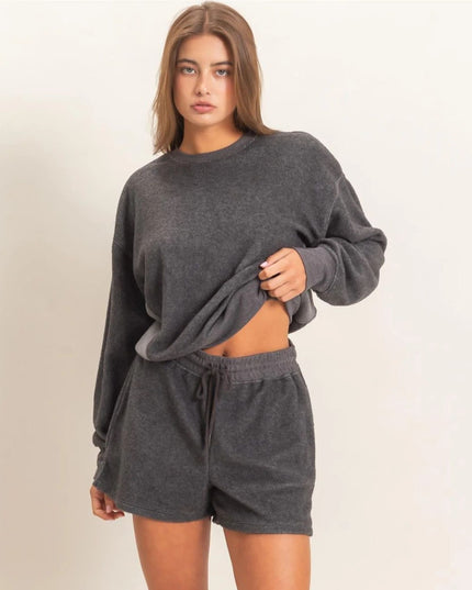 A woman modeling a dark gray two-piece loungewear set consisting of a cropped long-sleeve sweatshirt and matching drawstring shorts against a plain off-white background.