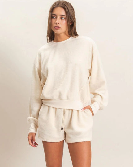A woman modeling a matching cream-colored lounge set consisting of a long-sleeve sweatshirt and drawstring shorts against a plain, light beige background.