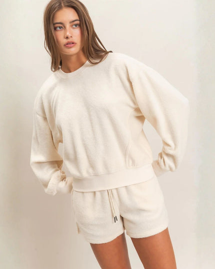 A woman modeling a matching cream-colored lounge set consisting of a cropped crewneck sweatshirt and drawstring shorts against a plain light beige background.