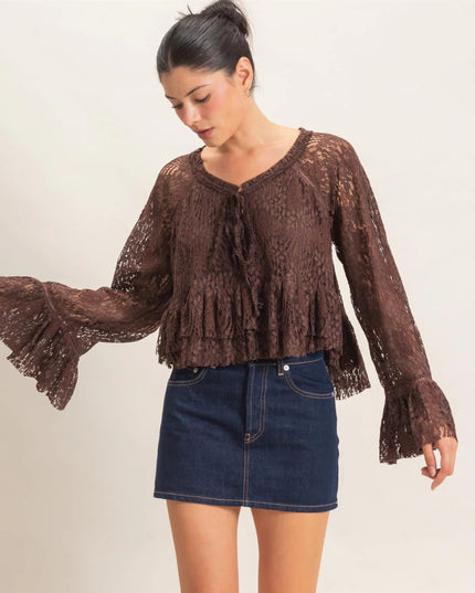 A woman wearing a brown lace long-sleeve blouse with a ruffled tie neckline and flared bell sleeves, paired with a dark blue denim mini skirt, against a plain light background.