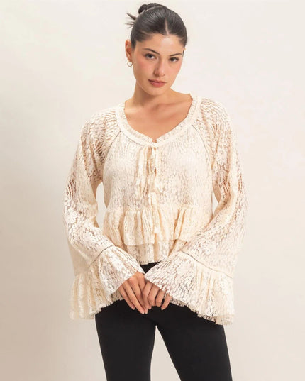 A woman modeling a cream-colored lace long-sleeve blouse with a ruffled tie neckline and bell sleeves, paired with black pants, against a plain light beige background.