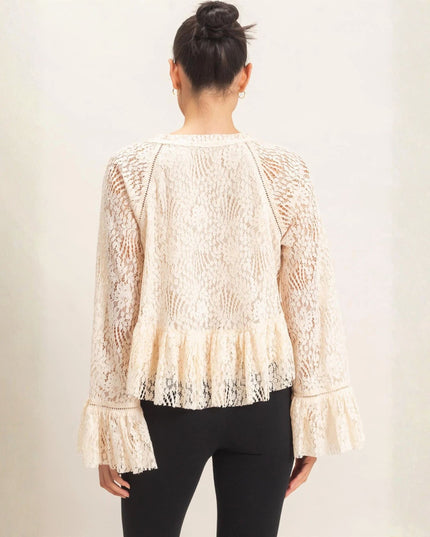 A woman viewed from the back wearing a cream lace long-sleeve top with a ruffled hem and cuffs, paired with black pants, against a plain light beige studio background.