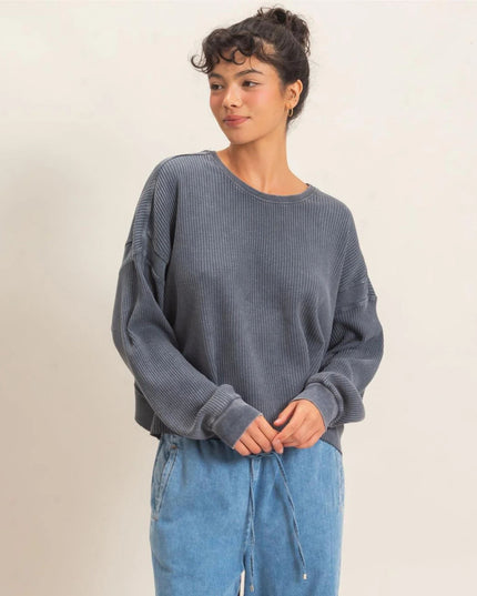 A woman wearing a dark slate blue-gray ribbed knit sweater and light blue jeans, standing against a plain off-white background.