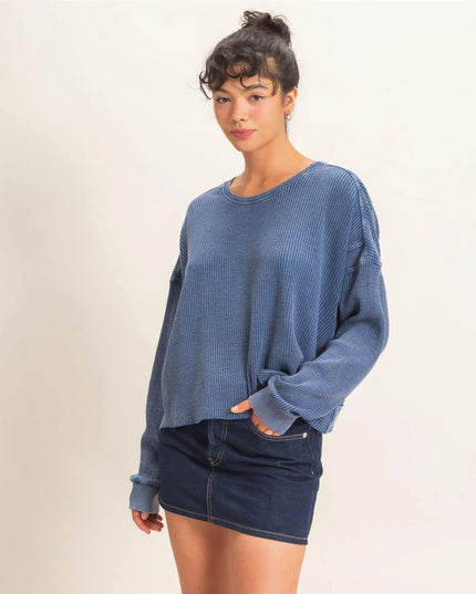 A woman modeling a blue ribbed knit long-sleeve crewneck sweater paired with a dark denim mini skirt, set against a plain off-white studio background.