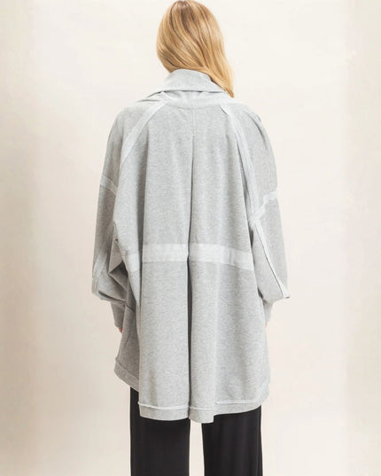 A woman shown from the back wearing a light gray heathered oversized cardigan with white contrast panels, paired with black pants, against a plain beige background.