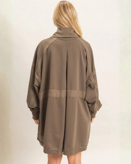 A woman viewed from the back wearing an oversized taupe-brown longline jacket with a mock neck and wide sleeves, set against a plain light beige wall.