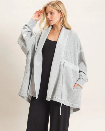 A woman modeling a light gray oversized zip-front hoodie with a shawl collar and drawstring, worn open over a black top and black pants, against a plain light beige studio background.