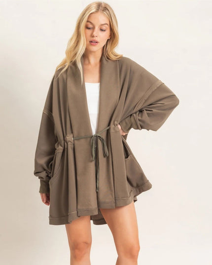 A woman modeling an oversized taupe-brown open-front cardigan with a drawstring waist, wide sleeves, and front pockets, worn over a white top against a plain light background.