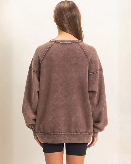 A woman viewed from the back wearing an oversized, washed brown crewneck sweatshirt and black bike shorts against a plain, light beige wall.