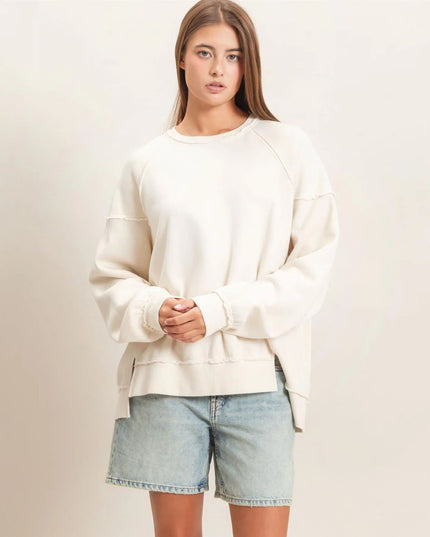 A woman modeling an oversized cream-colored crewneck sweatshirt with raglan sleeves, paired with light-wash denim shorts, against a plain beige studio background.