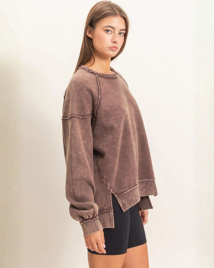 A woman modeling a brown washed, oversized long-sleeve sweatshirt with raw edges and elastic cuffs, paired with black bike shorts against a plain off-white background.