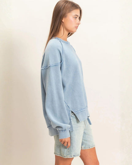 A woman wearing a light blue oversized crewneck sweatshirt and matching light blue denim shorts, shown from a side profile against a plain off-white wall.