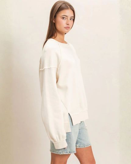 A woman modeling a cream-colored crewneck sweatshirt with a raw, frayed hem and cuffs, paired with light-wash denim shorts against a neutral cream background.