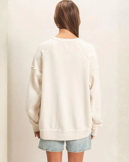 A woman viewed from behind wearing an oversized cream-colored crewneck sweatshirt and light-wash denim shorts, standing against a neutral beige wall.