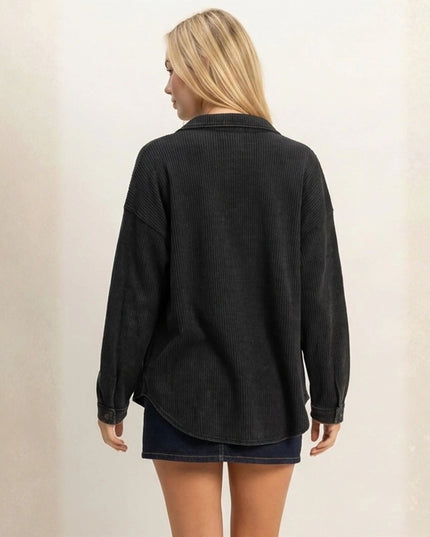A woman viewed from behind wearing a dark charcoal gray ribbed long-sleeve button-up shirt and a dark denim mini skirt, standing against a light beige textured wall.