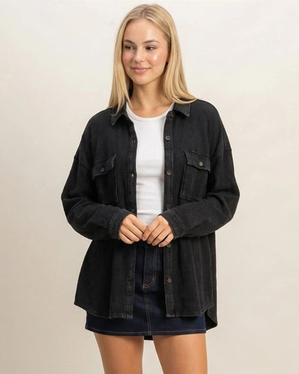 A blonde woman wearing a black corduroy button-up shirt jacket over a white t-shirt and a dark denim skirt, standing against a plain beige studio background.