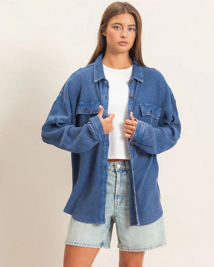 A woman modeling an oversized blue corduroy button-up shirt jacket over a white t-shirt and light-wash denim shorts, set against a plain off-white studio background.