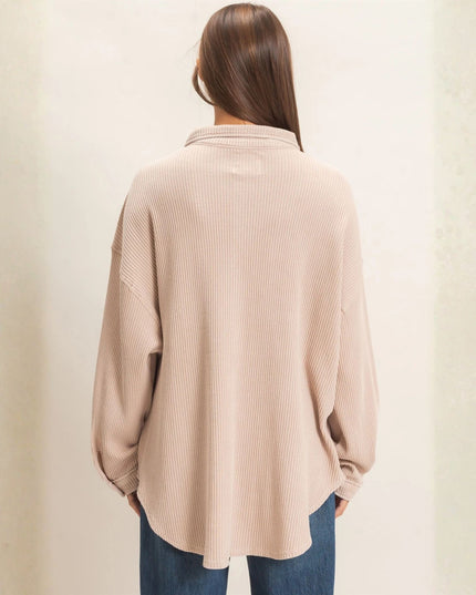 A woman viewed from behind wearing a beige ribbed knit long-sleeve shirt and blue jeans, standing against a textured off-white wall.