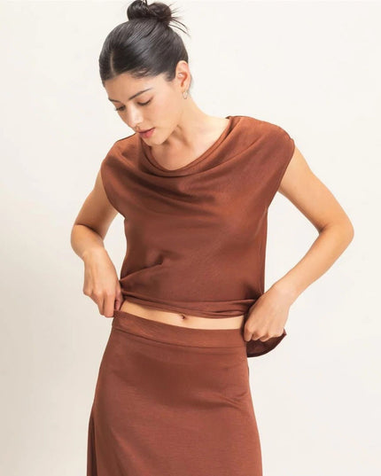 A woman modeling a matching rust-brown two-piece set consisting of a draped sleeveless crop top and a high-waisted skirt, against a plain off-white background.
