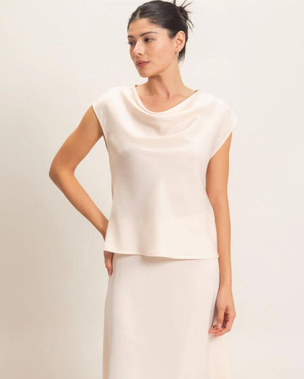 A woman modeling a light cream-colored cowl-neck top with cap sleeves and a matching high-waisted skirt against a plain light background.