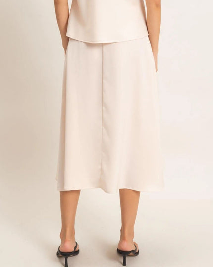 A model from the back wearing a light cream or off-white midi skirt with a draped, layered design and black slingback heels, set against a plain light beige studio background.