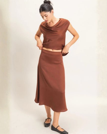 A woman modeling a rust-brown two-piece knit set consisting of a short-sleeve draped cowl-neck top and a matching midi skirt, paired with black Mary Jane flats against a plain off-white background.