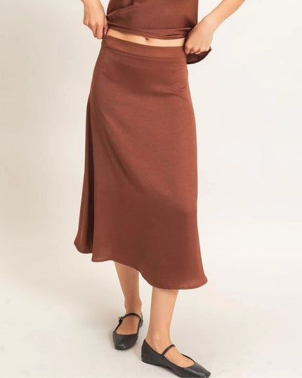 A person wearing a rust-brown midi skirt with a side slit and a matching brown top, paired with black flat shoes, against a plain light background.