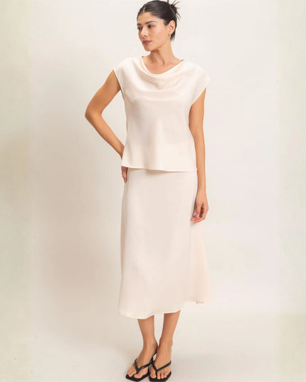 A woman modeling a two-piece cream-colored outfit consisting of a short-sleeve cowl-neck top and a matching midi skirt, paired with black sandals, against a plain off-white studio background.