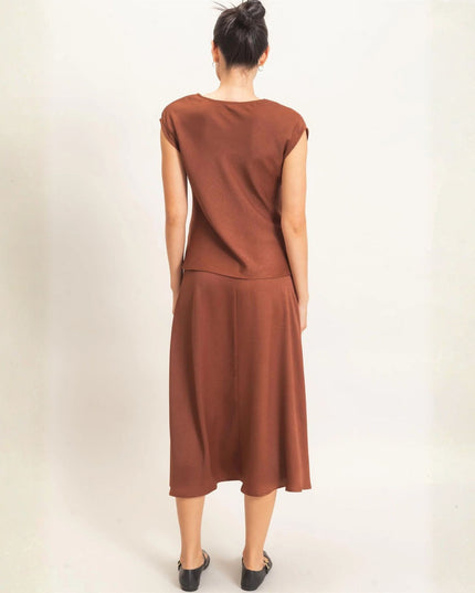A woman viewed from behind wearing a rust-brown sleeveless dress with a round neckline and a flowing midi skirt, standing against a plain off-white studio background.