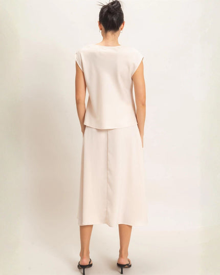 A woman viewed from behind wearing a light cream two-piece set consisting of a short-sleeve top and a midi skirt, paired with black heeled sandals against a plain off-white background.