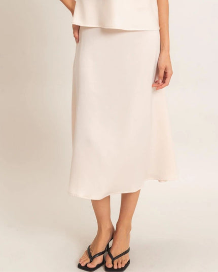 A model wearing a light cream or off-white midi skirt with a matching top and black thong sandals, shown from the waist down against a plain light beige studio background.