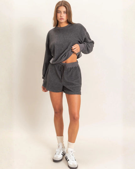 A woman modeling a dark gray two-piece lounge set consisting of a cropped long-sleeve sweatshirt and matching drawstring shorts, paired with white socks and white sneakers, against a plain off-white studio background.