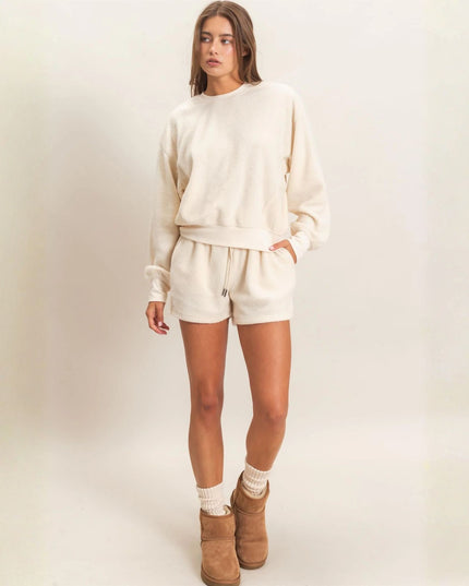 A woman modeling a matching cream-colored lounge set consisting of a long-sleeve sweatshirt and shorts, paired with brown suede boots and white socks, against a plain off-white studio background.