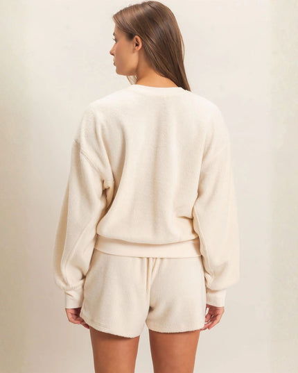 A woman shown from the back wearing a cream-colored crewneck sweatshirt and matching shorts against a neutral beige background.
