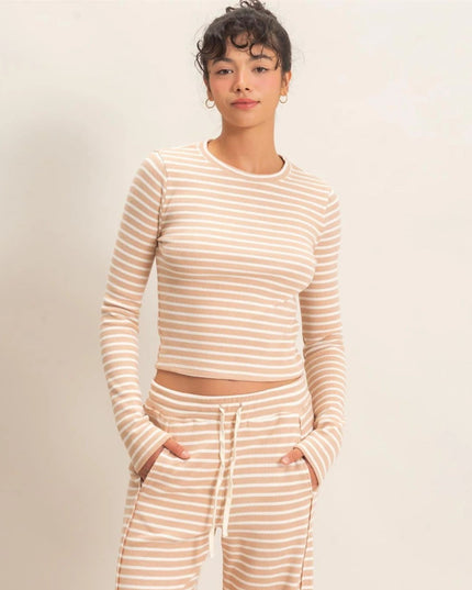 A woman modeling a matching beige and white striped long-sleeve crop top and drawstring sweatpants against a plain light beige studio background.