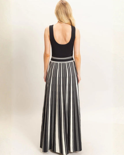 A woman viewed from the back wearing a sleeveless black top and a long black-and-white vertically striped pleated skirt, standing against a plain light background.