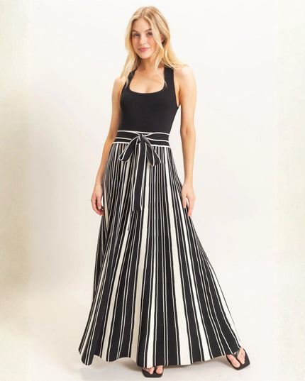 A woman modeling a sleeveless maxi dress with a solid black bodice and black and white vertical stripes on the skirt, featuring a matching tie belt at the waist.