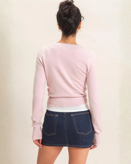 A woman shown from the back wearing a light pink long-sleeve knit sweater and a dark blue denim mini skirt, standing against a plain light beige wall.
