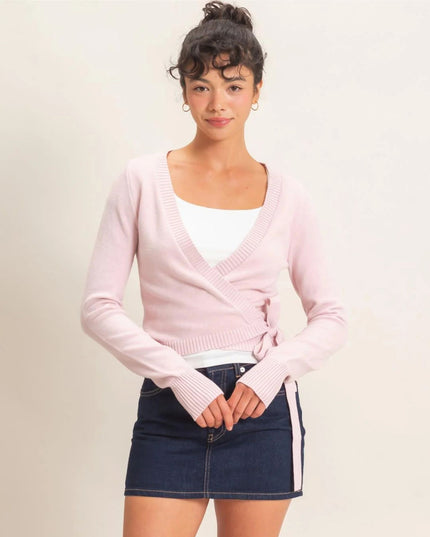 A woman modeling a light pink wrap cardigan over a white tank top and a dark denim mini skirt, photographed against a plain light beige studio background.