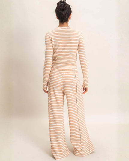 A woman viewed from behind wearing a beige and white horizontally striped long-sleeve jumpsuit with wide-leg pants, set against a plain off-white studio background.