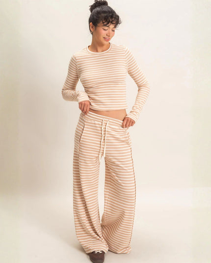 A woman modeling a matching two-piece loungewear set consisting of a beige and white horizontally striped long-sleeve crop top and matching wide-leg drawstring pants, set against a plain cream studio background.