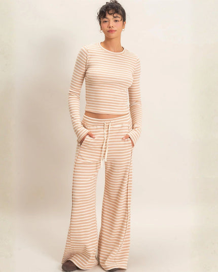 A woman modeling a matching two-piece loungewear set featuring a long-sleeve cropped top and wide-leg pants, both in a beige and white horizontal stripe pattern, set against a neutral cream background.
