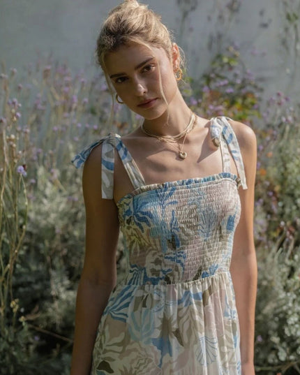 A woman wearing a smocked sleeveless dress with a blue, beige, and white botanical pattern, standing in a garden setting.