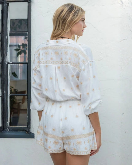 A woman shown from the back wearing a white embroidered romper with gold-toned floral and geometric patterns, standing against a white textured wall next to a black-framed window.