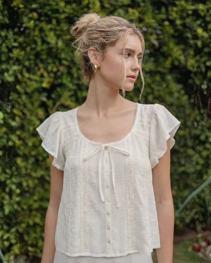 Woman wearing a white blouse with ruffled sleeves against a green hedge background