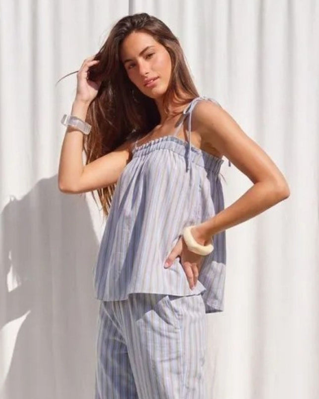 A woman modeling a light blue and white vertically striped two-piece pajama set, consisting of a tie-strap crop top and matching pants, posed against a white curtain backdrop.