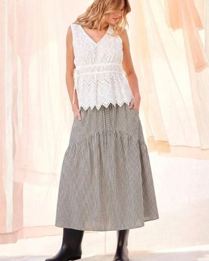 A woman wearing a sleeveless white lace peplum top and a gray and white vertically striped tiered midi skirt, paired with black boots, standing in front of a soft pink and white draped fabric backdrop.