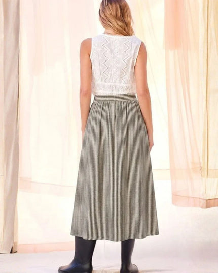 A woman shown from the back wearing a sleeveless white lace top and a high-waisted, floor-length pleated skirt with thin vertical stripes in gray and white. She is wearing black rain boots, standing in front of a soft, draped cream-colored curtain backdrop.